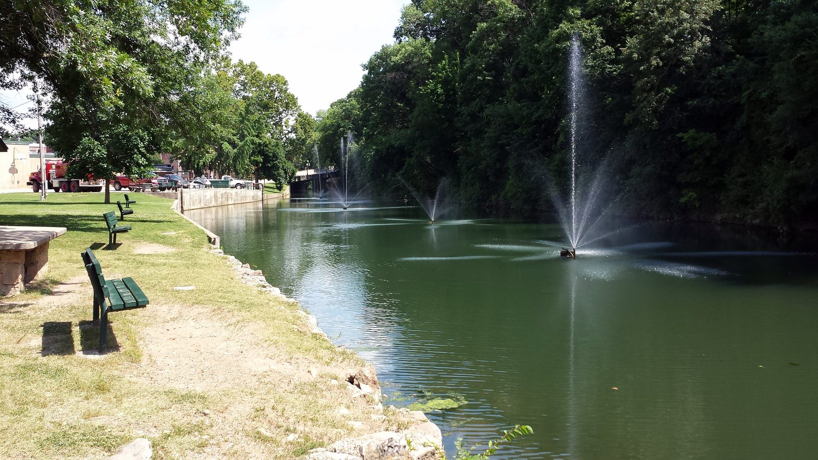 Sager Creek running through City Park in downtown Siloam Springs, with fountains in the water, green park benches along the bank and tall shade trees